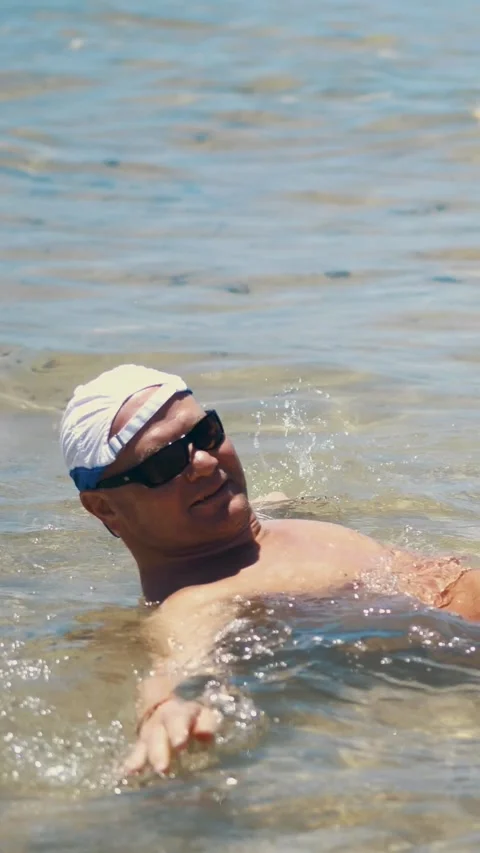 A man in swimming trunks, in sunglasses and a blazer lies in shallow water, Stock Footage 313484483