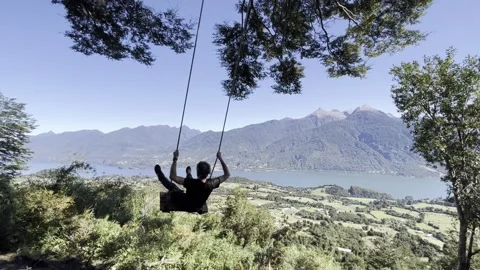 Man on Swing Overlooking Mountains and Lake in Cochamo Chile - 4K Video stock 318364260