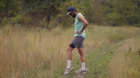 Man swinging leg forward in dynamic hamstring stretch on grassy trail outdoors Stock Footage 313951478