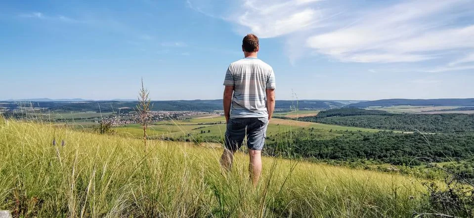 Man in a T-shirt and shorts looks down onto a town in a valley from a hill Stock Photos