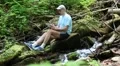 Man with tablet computer sits near small river in forest HD Footage