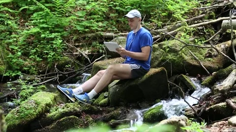 Man with tablet computer sits near small river in forest 스톡 동영상 56265236