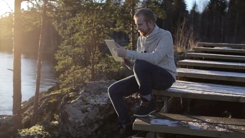 A man with a tablet sits on the steps of a rocky shore of the lake Stock Footage 82583044
