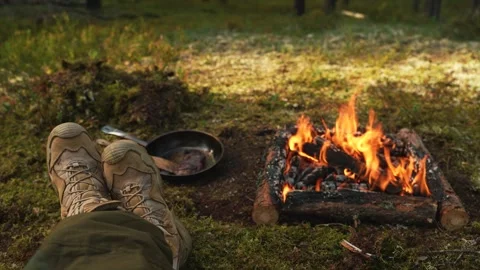 A man in tactical boots sits next to a fire in the woods. View of legs and fire. Video stock 249194994