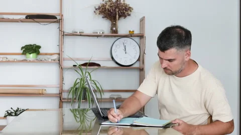 Man takes notes in a notebook while working on a laptop. Stock Footage 310254284