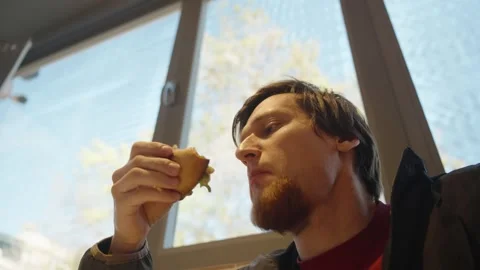 Man Taking a Bite of a Sandwich While Looking Up with a Large Window in the 스톡 동영상 303160429