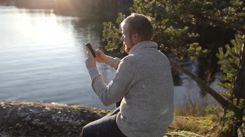 Man is taking pictures on the phone while sitting on the rocky shore of the lake Stock Footage 82583126