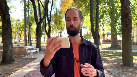 Man Talking and Presenting with a Smartphone in an Autumn Park Stock Footage 319820785