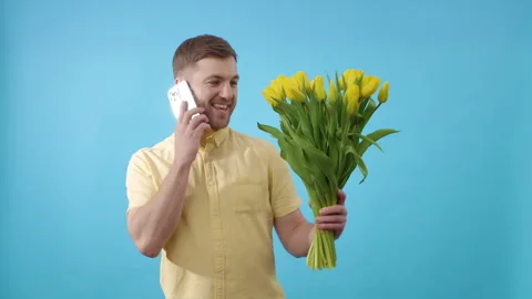 Man talking on cell phone holding bouquet of yellow tulips on blue background Video stock 266456912