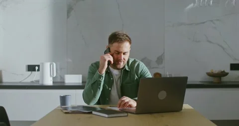 A man is talking on mobile phone, sitting at kitchen table in front of laptop Stock Footage