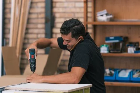 Man talking with a mobile while drilling a piece of furniture in a workshop Stock Photos