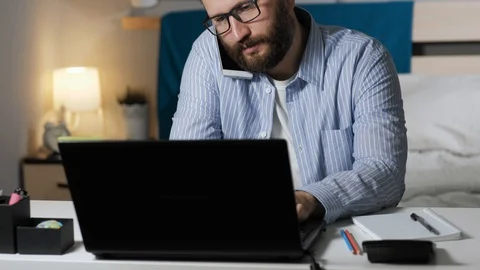 Man is talking on phone and working on computer. Bearded guy sits at desk in Stock Footage 129375455