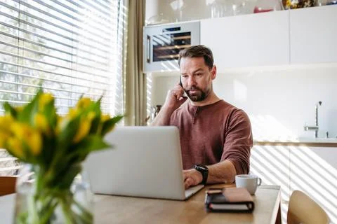 Man talking on phone with serious expression while working remotely on laptop Stock Photos