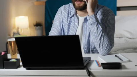Man talking on phone working and drinking beer. Bearded guy sits at desk in Stock Footage 129378900