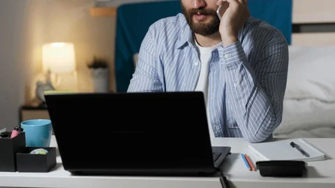 Man talking on phone working and drinking coffee. Bearded guy sits at desk in Stock Footage 129379276