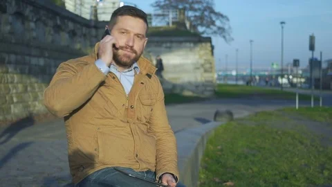Man talking by smartphone, sitting on wall at boulevard next to river  Vídeo Stock 72775444