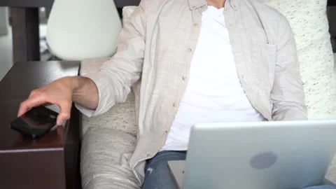 Man talking on smartphone while sitting on couch with laptop, working remotely Stock Footage 154796371
