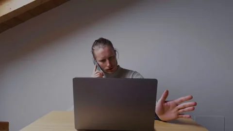 Man talking on smartphone while working on laptop from home 스톡 동영상 334735955