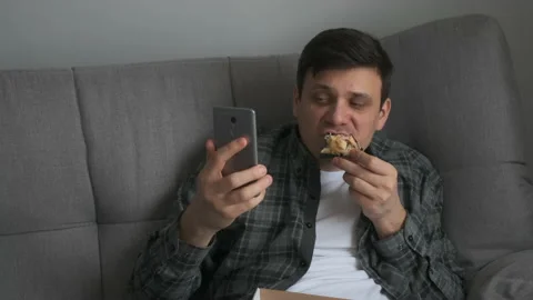 Man talking video chat on smartphone and eating pizza sitting on sofa at home. Video stock 158804862