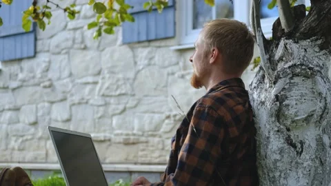 Man talking on a video link on a laptop while sitting under a tree Stock Footage 200825330
