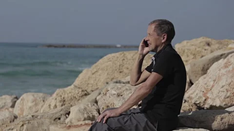 Man talks to a cell phone sitting on the stones near sea Stock Footage 57371050