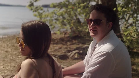 Man talks to his friends while sitting on a blanket next to a lake 스톡 동영상 116765558