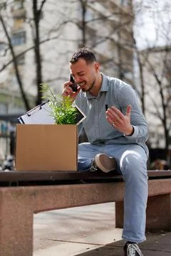 Man talks on phone while sitting on bench with a box and a plant outside in the Stock Photos