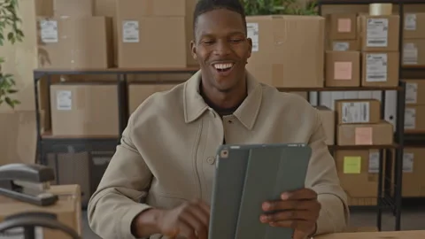 Man in tan jacket holding tablet waves hand and smiles beside stacked parce.. Stock Footage 337434931