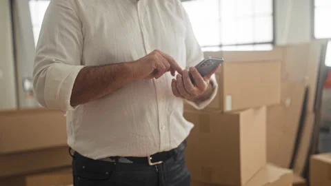 Man tapping smartphone with hands inside building among stacked boxes and a.. Stock Footage 339200895
