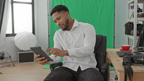 Man tapping tablet in studio beside camera gear and green screen, seated in.. Stock Footage 337439876