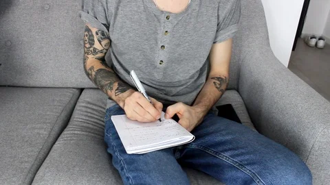 Man in tattoos writing on a notebookon the couch Vídeos de archivo 86572333
