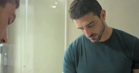 Man in teal tee with bracelet, raising gaze at mirror, tilting dropper, rubbing Stock Footage 344105599
