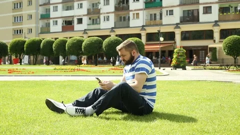 Man texting on smartphone while sitting on the grass in the park Stock Footage
