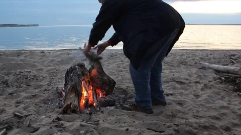 Man throwing firewood at a bonfire on the river bank at sunset Stock Footage 81008567