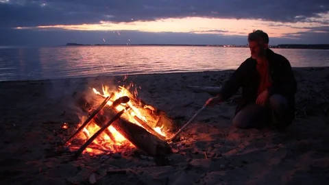 Man is throwing firewood at a fire on the river bank at sunset Stock Footage 81008571