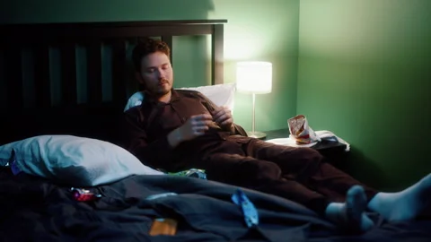 Man throwing smartphone in irritation and taking chips in bed at night Video stock 319443262