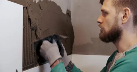 Man tiler puts solution on wall with ribbed spatula, smooths it with even layer Stock Footage 130777425