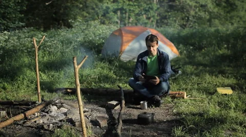 A man touch a tablet near the campfire and tents. In the forest morning Stock Footage 64313586