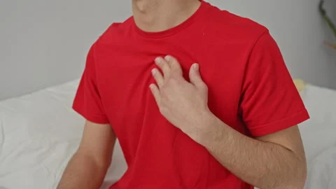 Man touching chest with hand while seated on bed in bedroom, red tshirt vis.. Stock Footage 337376405