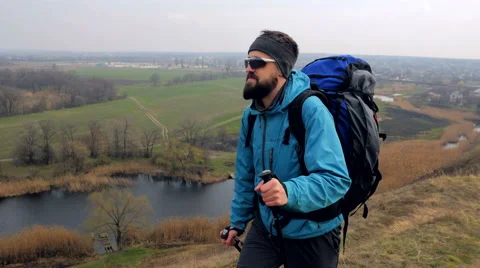 Man tourist in a blue jacket with a backpack and trekking poles walks outdoor Stock Footage 62398300