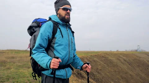 Man tourist in a blue jacket with a backpack and trekking poles walks outdoor Stock Footage 62398574
