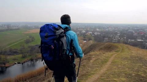 Man tourist in a blue jacket with a backpack and trekking poles walks outdoor Stock Footage 62398995