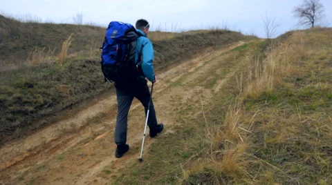 Man tourist in a blue jacket with a backpack and trekking poles walks outdoor Stock Footage 62399366