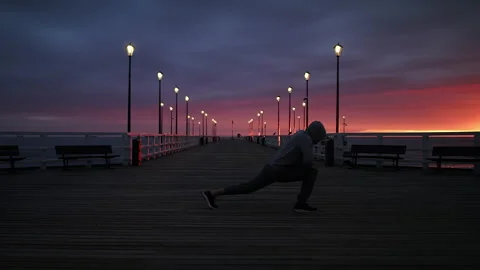 Man In Tracksuit and Protective Face Mask Stretching on a Seaside Pier at Dusk - Stock Footage 130733710