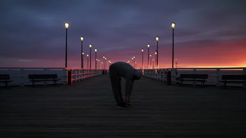 Man In Tracksuit and Protective Face Mask Stretching on a Seaside Pier at Dusk - Stock Footage 130737072