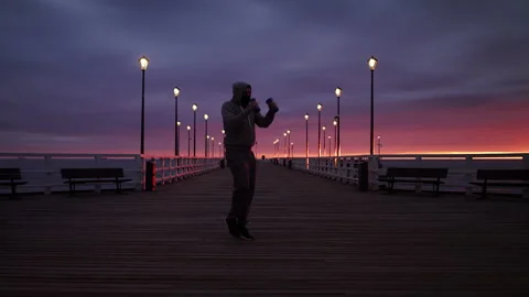 Man In Tracksuit Training with Weights on a Seaside Pier at Dusk - Wide Shot Stock Footage 130729917