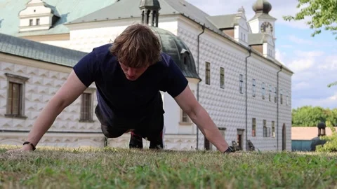 Man trains outdoors doing one arm push up on grass near historic building showin Vídeos de archivo 315413815