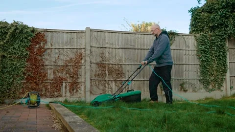 Man trimming backyard grass with electric lawnmower near fence. Male mowing Stock Footage 306476783