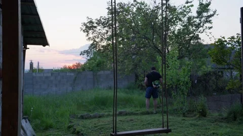 Man Trimming Grass in Backyard at Sunset With Swing in Foreground Vídeo Stock 341766967