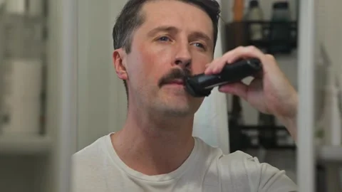 A man trims his mustache using an electric trimmer while standing in a bathroom. 库存影片 337111599